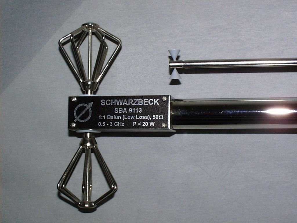 Figure 5 — A small biconical microwave antenna with its specification plate legible: SBA 9113, 1:1 balun, 50 ohms, 0.5 to 3 GHz, power under 20 watts. Source: Wikimedia Commons, Schwarzbeck Mess-Elektronik, C…