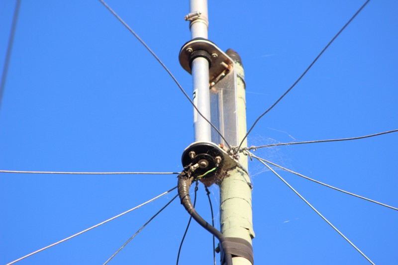Figure 3 — Close-up of a trap junction and guy-wire attachment point on a field-installed Hustler-family trap vertical, showing the aluminum trap enclosure and the multiple tensioned support lines a tall mult…