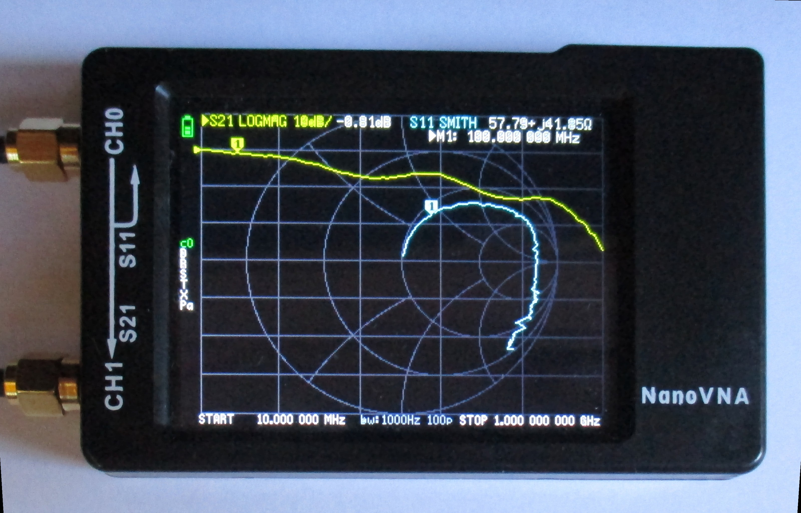 Figure 4 — A NanoVNA displaying a live S11 Smith-chart and S21 LOGMAG trace — the same CH0/S11 display convention the sweep-and-trim workflow below reads. Photo: File:NanoVNA S11 S21.jpg by Martin Homuth-Rose…