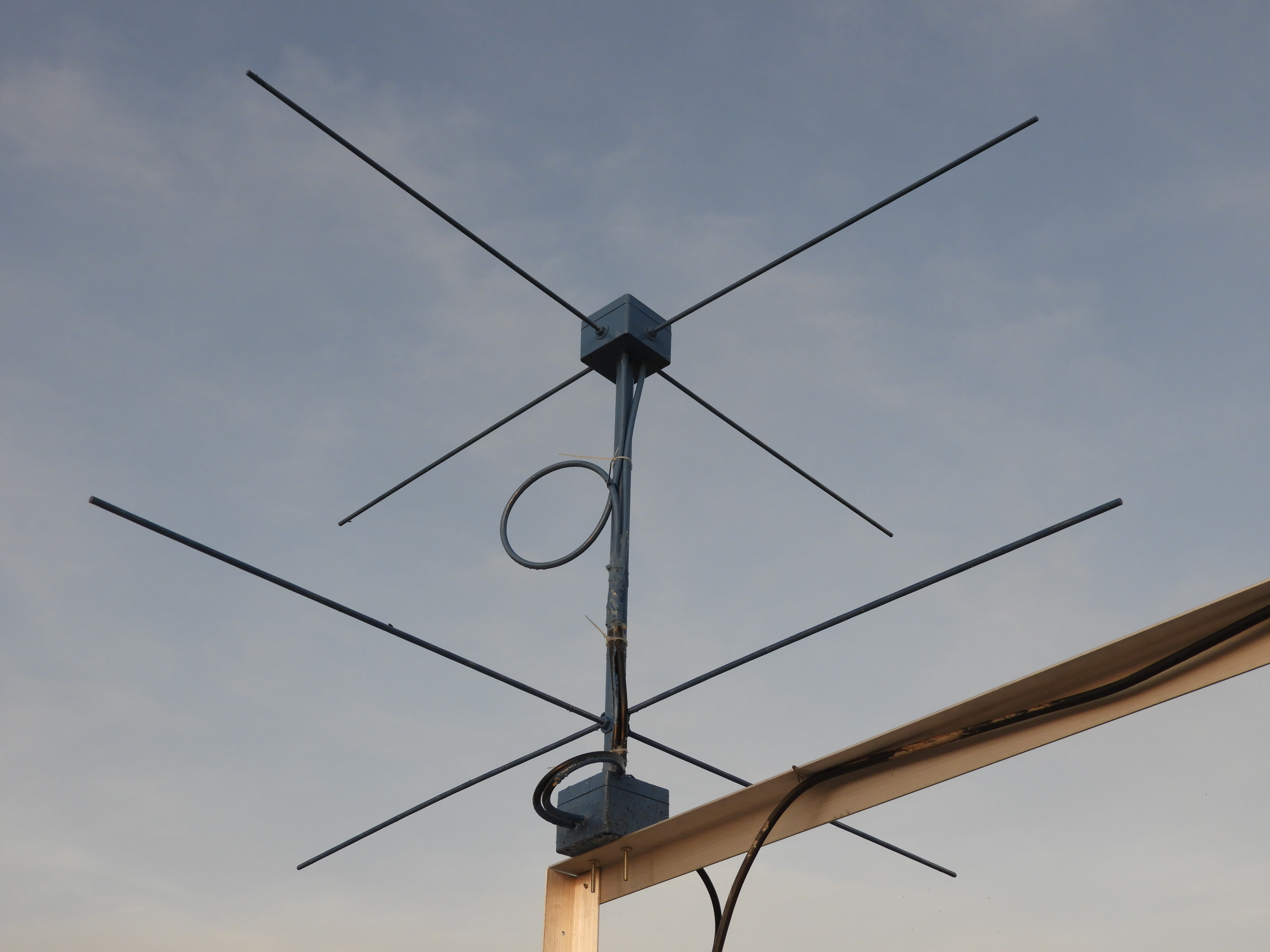 Figure 6 — A home-built 137 MHz turnstile: threaded-rod elements in two die-cast boxes on a timber post, with a coiled-coax common-mode choke visible at each feed. Photograph by Dilshan Jayakody, CC BY-SA 2.0…